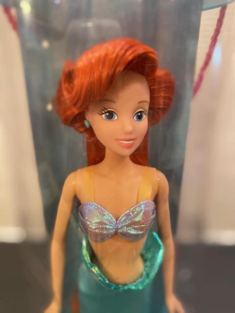 May include: A doll of a red-haired mermaid with blue eyes and a shimmering seashell top. The doll has a teal tail and is a popular character from a children's animated movie.