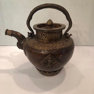 Könnte beinhalten: Eine bronzefarbene Teekanne mit aufwendigen geprägten Mustern und einem gebogenen Griff. Die Teekanne hat einen Ausguss, einen Deckel mit Knauf und dekorative Elemente um den Boden und den Körper. Das Design deutet auf einen Vintage- oder Antik-Stil hin.