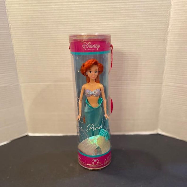 May include: Ariel doll in a clear plastic tube. The doll has red hair, a silver top, and a teal mermaid tail. The tube has a pink top with the Disney Princess logo and a red handle. The doll is approximately 12 inches tall.