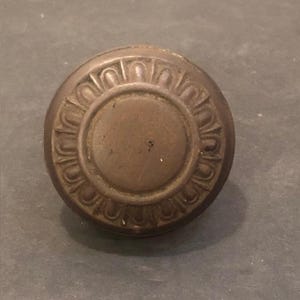 May include: A round, dark bronze-coloured metal door knob featuring a smooth, flat circular centre surrounded by a raised, repeating decorative border pattern of vertical, arched segments that extend to the outer edge of the knob's face.