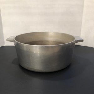 May include: A round, silver-coloured metal cooking pot with a wide, open top and two integrated side handles. The exterior and interior surfaces have a brushed metallic finish, showing a simple, functional design for stovetop food preparation.