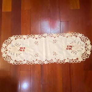 Può includere: Runner da tavolo color crema con bordo in pizzo smerlato. Il runner presenta ricami floreali e a nastro in rosso e bianco. Il runner è posto su una superficie di legno.