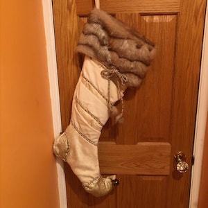 May include: A cream-colored Christmas stocking with a faux fur cuff and gold accents. The stocking is hanging on a wooden door. The stocking has a decorative gold trim and a gold toe.