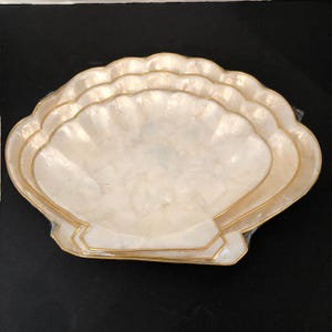 Puede incluir: Un juego de cuatro bandejas para servir con forma de concha. Las bandejas tienen un interior blanco nacarado y un borde dorado. Las bandejas están apiladas una encima de la otra y el fondo es negro.