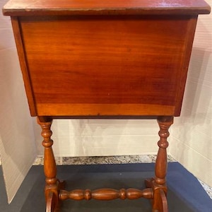 May include: A medium-brown wooden sewing stand featuring a rectangular storage compartment with a hinged lid and a handle cutout at the top. The unit is supported by two turned vertical legs connected by a horizontal stretcher bar at the base.