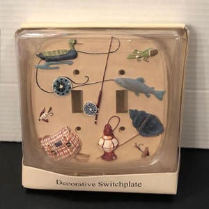 May include: A light-colored double toggle switchplate featuring raised fishing-themed motifs including a duck, a fish, a fishing rod, a reel, a net, a lantern, and a plaid bag. The item is encased in clear plastic packaging labeled Decorative Switchplate.