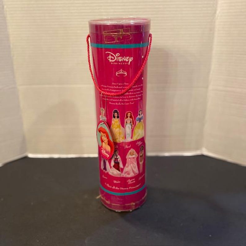 May include: A cylindrical, pink Disney Princess collectible container with a red handle. The container features images of Disney princesses and the Disney logo. A tag with Ariel's image hangs from the container.