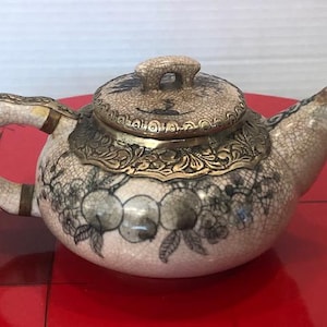 May include: A small, antique teapot with a cream-colored, crackled finish. The teapot features a decorative gold-colored band around the lid and base, and a handle and spout. The body of the teapot is decorated with a floral pattern in shades of gray and black.