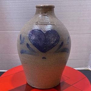 Puede incluir: Jarra de gres con un diseño de corazón azul oscuro y detalles florales. La jarra es de color beige neutro, con un cuello estrecho y un cuerpo redondeado. Tiene una estética vintage y rústica.