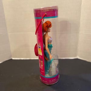 May include: A doll of a mermaid with red hair, a teal tail, and a seashell top, packaged in a clear plastic tube with a pink and teal label. The doll is approximately 12 inches tall.