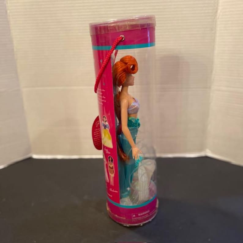 May include: A doll of a mermaid with red hair, a teal tail, and a seashell top, packaged in a clear plastic tube with a pink and teal label. The doll is approximately 12 inches tall.