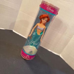 May include: Ariel doll in a clear plastic tube. The doll has red hair, a seashell top, and a teal tail. The tube has pink and teal accents and the Disney Princess logo.