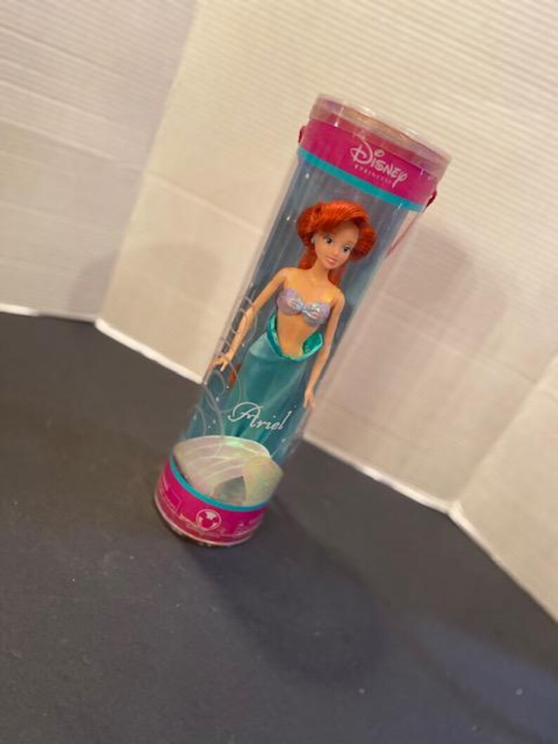 May include: Ariel doll in a clear plastic tube. The doll has red hair, a seashell top, and a teal tail. The tube has pink and teal accents and the Disney Princess logo.
