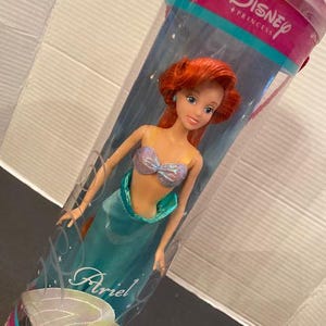 May include: Ariel doll from Disney Princess, in a clear plastic tube. The doll has red hair, blue eyes, and a teal mermaid tail. The tube has the Disney Princess logo and the name "Ariel" on it.