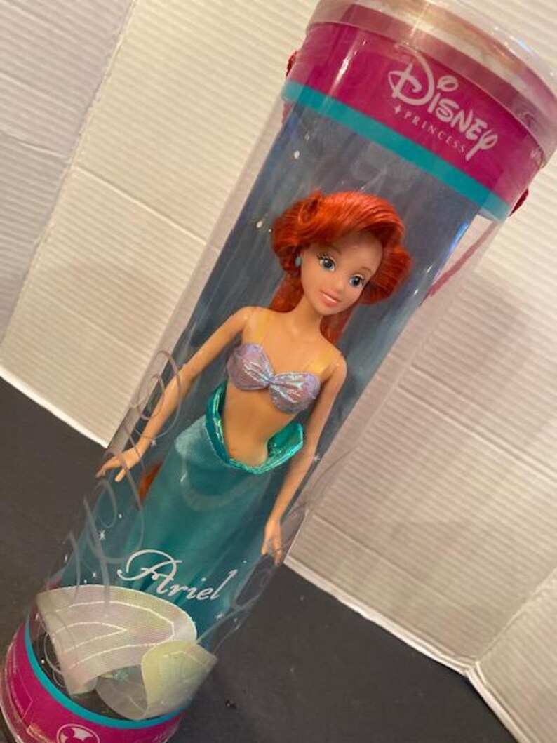 May include: Ariel doll from Disney Princess, in a clear plastic tube. The doll has red hair, blue eyes, and a teal mermaid tail. The tube has the Disney Princess logo and the name "Ariel" on it.