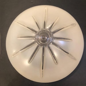 May include: A circular, frosted white glass ceiling light shade featuring a clear, raised starburst pattern radiating from a central circular point. The design consists of twelve distinct, tapered rays extending outward toward the edge.