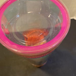 May include: A clear plastic tumbler with a bright pink rim. The tumbler appears to be partially filled with a reddish-orange substance. The image is taken from a top-down perspective, showing the inside of the cup.