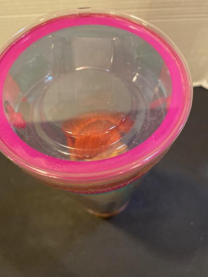 May include: A clear plastic tumbler with a bright pink rim. The tumbler appears to be partially filled with a reddish-orange substance. The image is taken from a top-down perspective, showing the inside of the cup.