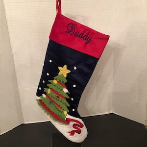 May include: A Christmas stocking with a red cuff embroidered with the word "Daddy" in cursive. The navy blue body features a felt Christmas tree with a gold star, red garland, and white snow. The toe is white with a red ribbon.