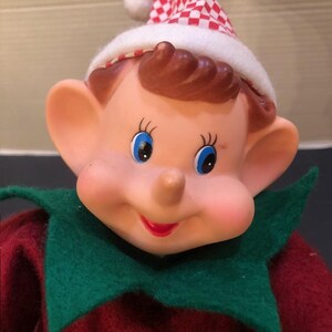 May include: A vintage elf doll with a rosy complexion, blue eyes, and a red smiling mouth. The elf wears a red and green outfit with a checkered hat. The doll has large ears and brown hair.