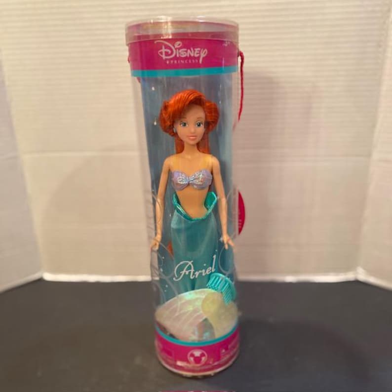May include: A Disney Princess Ariel doll in a clear plastic tube. The doll has red hair, a blue and silver top, and a teal tail. The tube has a pink top and bottom with the Disney Princess logo.