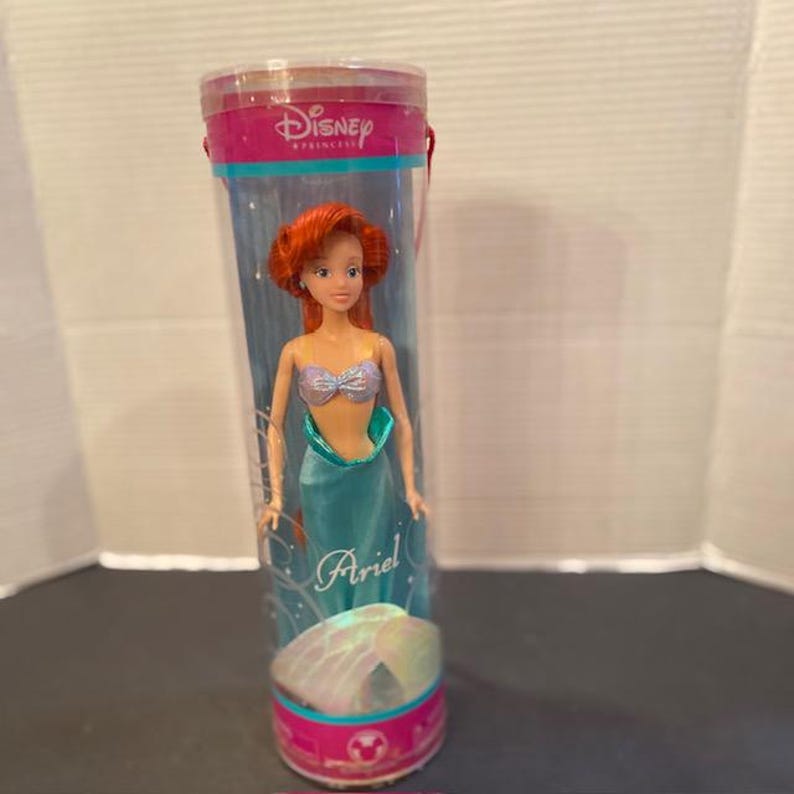 May include: Ariel doll from Disney's The Little Mermaid, in a clear plastic tube. The doll has red hair, a silver top, and a teal mermaid tail. The tube has a pink top with the Disney Princess logo and the name "Ariel".
