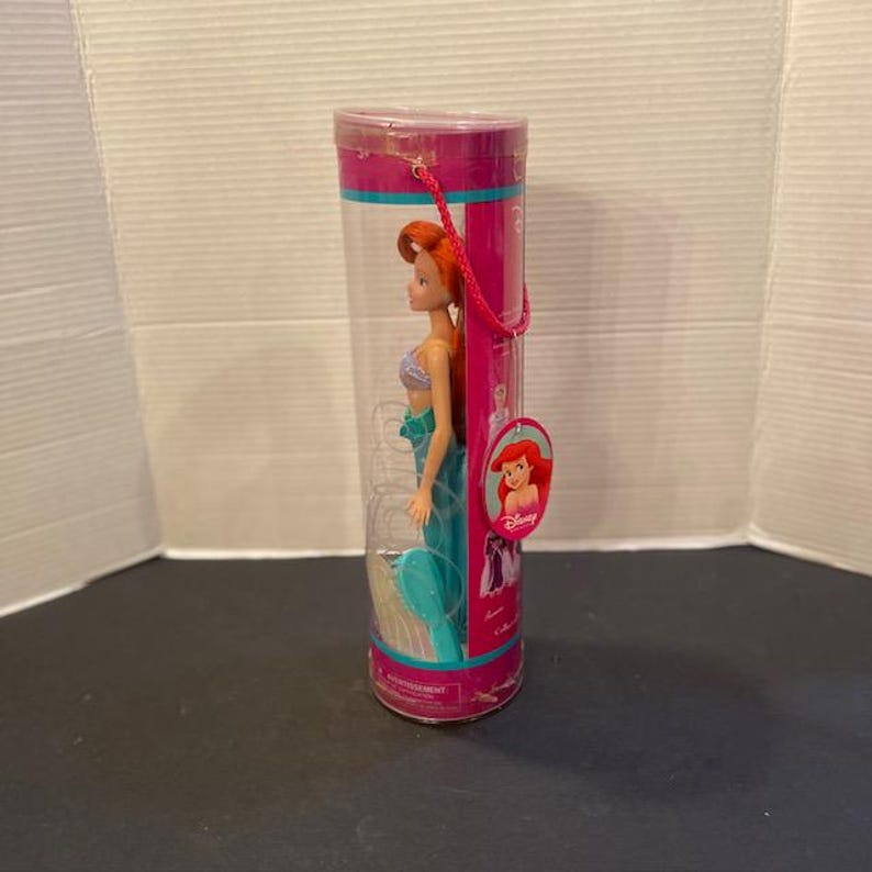 May include: Ariel doll in a clear plastic cylinder. The doll has red hair, a purple top, and a teal tail. The cylinder has pink and teal accents and a pink handle. A Disney tag with Ariel's image is attached.