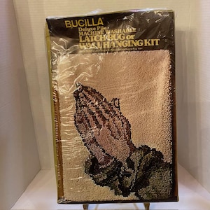 Könnte beinhalten: Ein Bucilla Deluxe Pile Latch Rug oder Wandbehang Kit. Das Set zeigt ein Design von betenden Händen in Braun-, Beige- und Grüntönen. Die Verpackung enthält den Text "MACHINE WASHABLE".