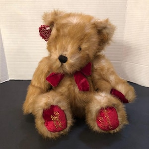 May include: A plush teddy bear with light brown fur, a black nose, and a red velvet bow. The bear has red velvet accents on its paws, with gold embroidered text. A red tag with "100th Anniversary" is attached to its ear.