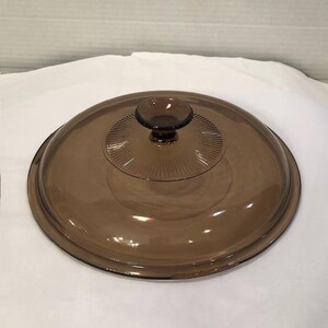 May include: A round, brown glass lid with a central knob. The lid has a slightly domed shape and a series of radial lines emanating from the knob. The knob is a darker shade of brown. This is a kitchenware item.