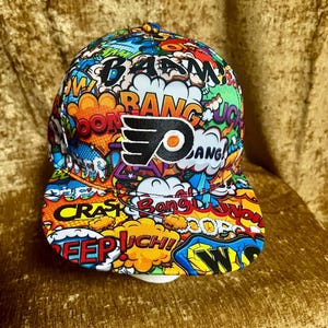May include: A baseball cap featuring a vibrant comic book-style print with colorful speech bubbles and sound effect text like BAAM, BANG, and CRASH. The front center displays the black and orange Philadelphia Flyers logo on a white background.