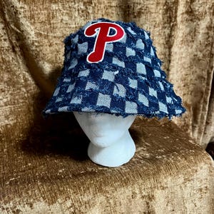 May include: A bucket hat made of blue and light blue checkered denim fabric with a frayed, distressed texture. The front of the hat features a large, embroidered red letter P with a white outline, centered on the crown of the hat.