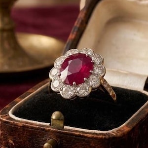 May include: A vintage ruby and diamond ring displayed in a leather jewellery box. The ring has a large, oval-cut ruby centre stone, encircled by a halo of smaller diamonds. The ring is set in a gold band. The box has a black velvet lining.