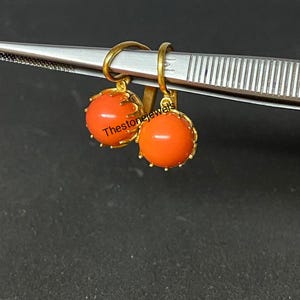 Könnte beinhalten: Ein Paar goldfarbene Ohrringe mit runden, orangeroten Edelsteinen. Die Edelsteine sind in einer dekorativen Goldfassung gefasst. Die Ohrringe werden von einem silbernen Werkzeug gehalten. Der Text "Thestonejewels" ist sichtbar.