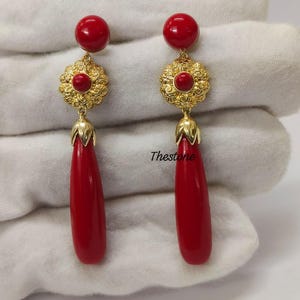 Könnte beinhalten: Ein Paar Ohrhänger mit einer runden roten Perle oben, einer goldenen, floralen Filigranscheibe mit einer kleinen roten Perle in der Mitte und einem langen, glatten, tropfenförmigen roten Anhänger an der Basis. Der Text Thestone ist sichtbar.