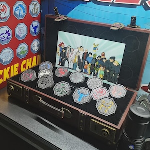 Può includere: Una valigia marrone in stile vintage aperta per rivelare una collezione di gettoni ottagonali color argento con vari disegni di animali. I gettoni sono esposti su un interno in velluto nero, con un'illustrazione colorata sullo sfondo. Le parole "ADVENTURES" e "JACKIE CHAN" sono visibili.