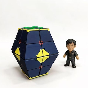 Könnte beinhalten: Ein geometrisches Puzzlespielzeug mit marineblauem und gelbem Design, akzentuiert mit grünen und roten Details. Eine kleine Figur eines Mannes in schwarzer Uniform steht daneben. Das Puzzle hat eine komplexe, facettenreiche Form.