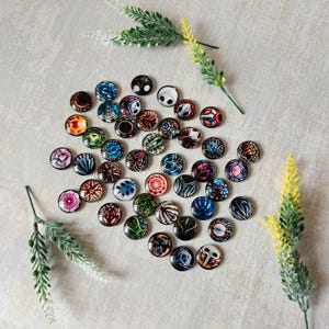 Op de afbeelding: Een verzameling ronde, kleurrijke knoopspelden met diverse ontwerpen, waaronder geometrische patronen, bloemmotieven en abstracte kunst. De spelden zijn verspreid op een lichtgrijze ondergrond, met takjes groen en gele bloemen.