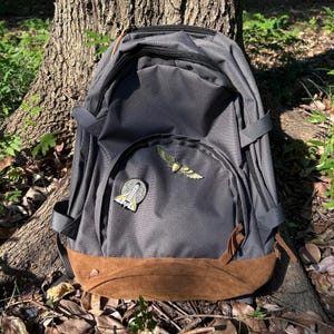 Może przedstawiać: Antracytowy szary plecak z brązowym panelem dolnym zamszowym. Plecak ma dwie ozdobne przypinki: jedną z uskrzydlonym przedmiotem i drugą z rakietą. Plecak opiera się o pień drzewa.