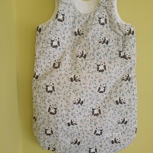 May include: A sleeveless baby sleeping bag featuring a repeating pattern of black and white pandas amidst small blue leaves on a light background. The garment includes two teal snap buttons at the shoulders and a square pocket on the front right side.