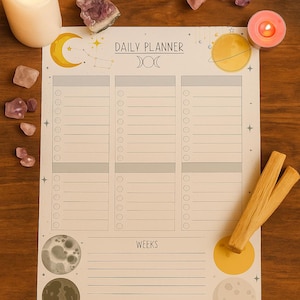 Puede incluir: Un planificador diario con ilustraciones celestiales, que incluyen lunas, estrellas y planetas. El planificador presenta secciones para la programación diaria, con la palabra "WEEKS" en la parte inferior. También hay una vela, cristales y palos de palo santo.