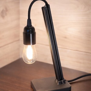 Puede incluir: Una taza de cerámica blanca con un asa negra y el texto "But First, Coffee" en negrita negra. La taza se encuentra sobre un fondo borroso, sugiriendo un ambiente acogedor.