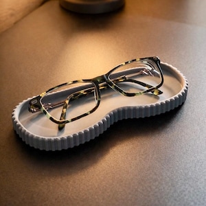 May include: A pair of eyeglasses with a tortoiseshell pattern rests on a gray, wavy-edged tray. The tray is on a dark surface, with a lamp in the background. The glasses have a rectangular frame.