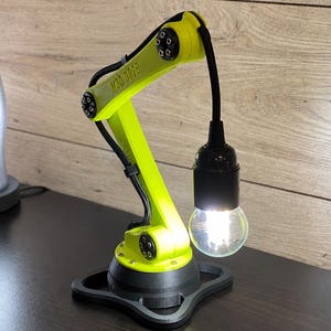 May include: A neon yellow robotic arm desk lamp with a black base and black joints. The arm features the text M3D 0828 printed on the side. A black light socket hangs from the end of the arm, holding a glowing light bulb with a visible filament.