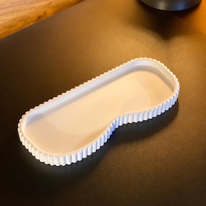 May include: A white, kidney-shaped tray with a scalloped edge. The tray is made of a solid, opaque material and sits on a dark surface. The tray's design is simple and elegant, suitable for various uses.
