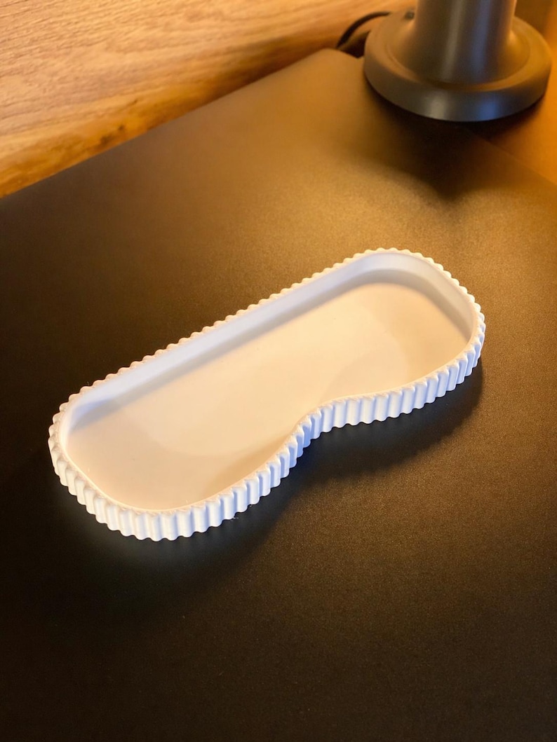 May include: A white, kidney-shaped tray with a scalloped edge. The tray is made of a solid, opaque material and sits on a dark surface. The tray's design is simple and elegant, suitable for various uses.