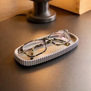May include: Eyeglasses with a tortoiseshell design are placed in a white, rectangular tray with a ridged edge. The tray is on a dark surface, with a lamp and wooden box in the background.