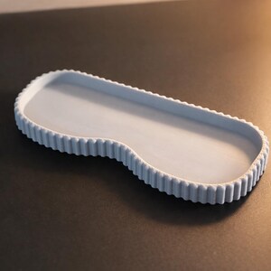 May include: A light blue, oblong decorative tray with a scalloped edge. The tray has a smooth interior and a wavy, asymmetrical design. The tray is made of a solid material and is suitable for displaying small items.