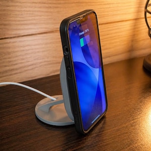 May include: A black smartphone in a case is charging on a grey wireless charging stand. The phone screen displays a blue background with a charging indicator showing 30%. A white charging cable is connected to the stand.