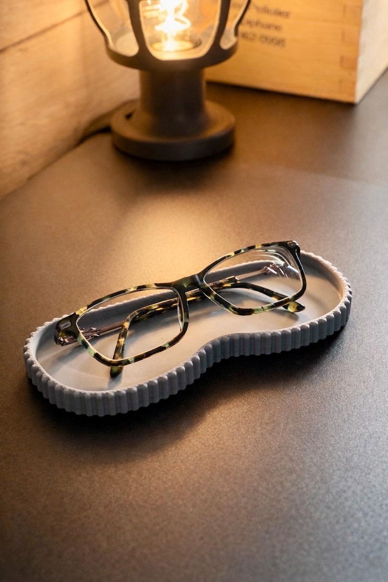 May include: A pair of eyeglasses with a tortoiseshell pattern rests on a gray, wavy-edged tray. The tray is on a dark surface, with a lamp in the background. The glasses have a rectangular frame.